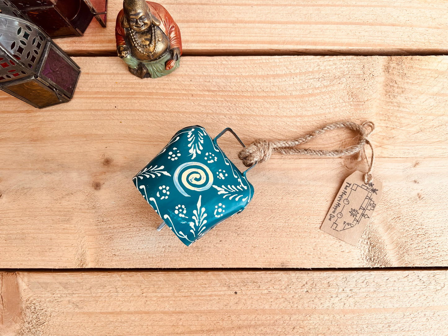 Hand painted traditional Indian hanging cow bell wind chime in blue colour with white floral feather painted detail and metallic swirl hand painted pattern handmade fair trade & ethically sourced with rustic jute twine hanging