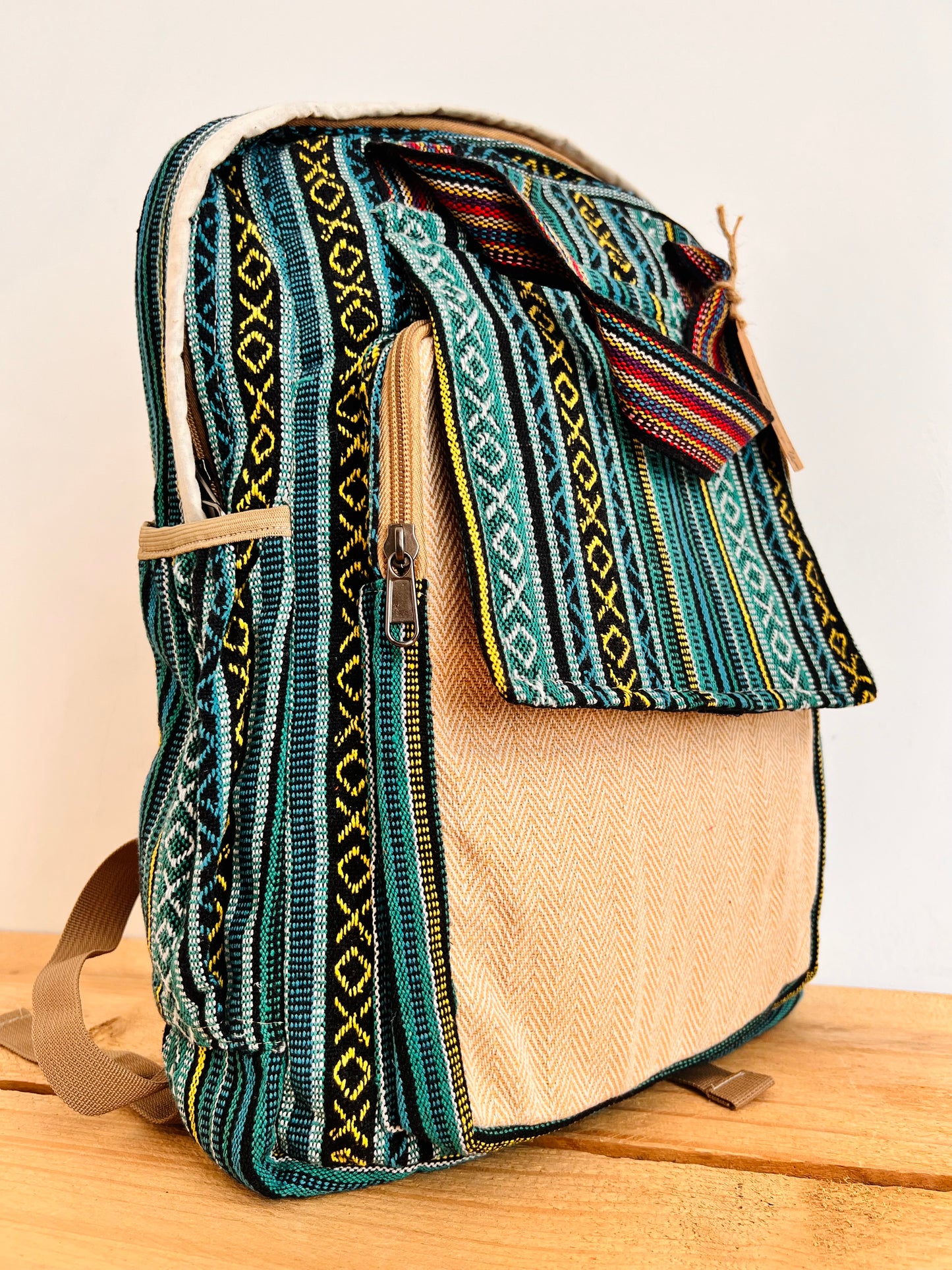 A woven backpack with a teal, black, and beige geometric pattern, featuring a tan canvas front pocket and a matching fabric top flap.