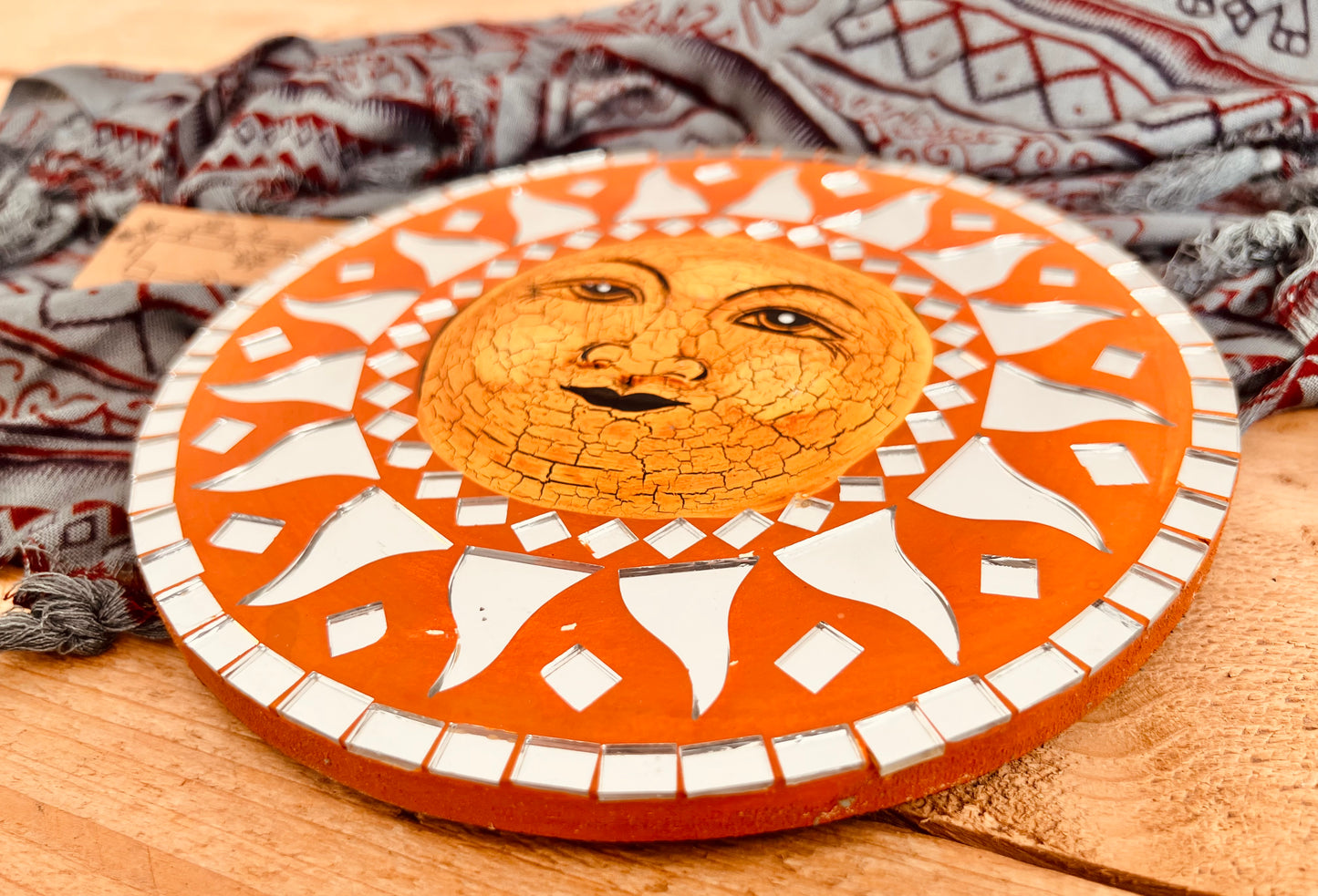 A round wooden sun wall plaque with a mosaic design, featuring a cracked-glass style sun face in the centre and mirror tile mosaic geometric rays radiating outward on an orange-brown background.